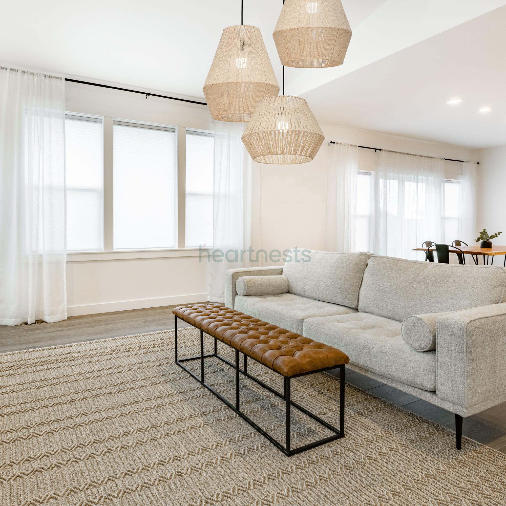 A Cluster of 1 Heartnests's Long Diamond Jute Hanging Light and 2 of Short Diamonds Jute Pendant Lights with lights on are hung above a wooden bench and grey lounge in a contemporary living room setting