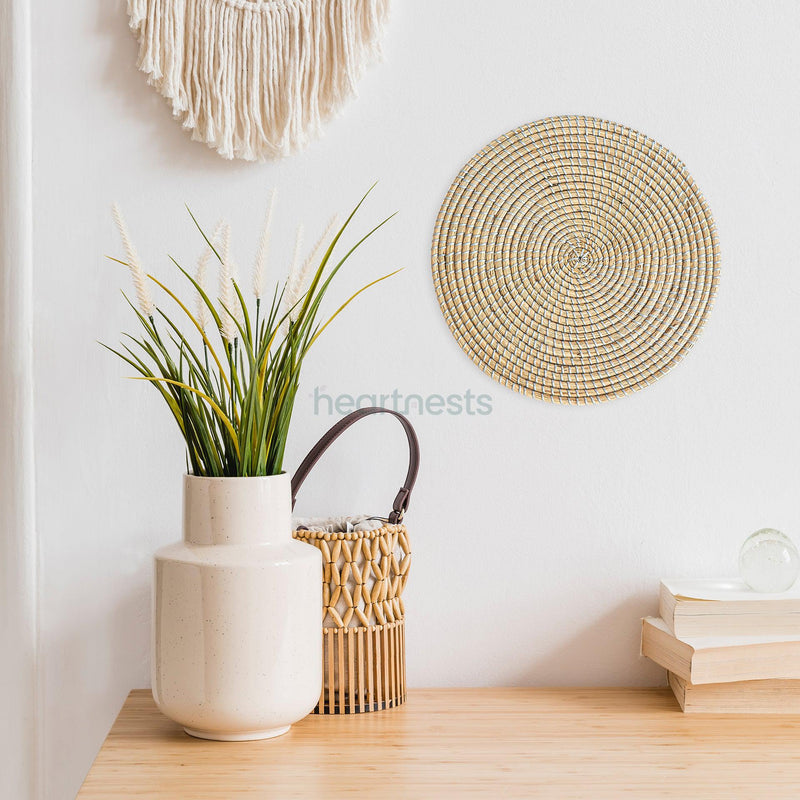 Heartnests' Ricey seagrass wall decor is hung on a while wall above a wooden table, a stack of books, a white flower vase and a rattan bag