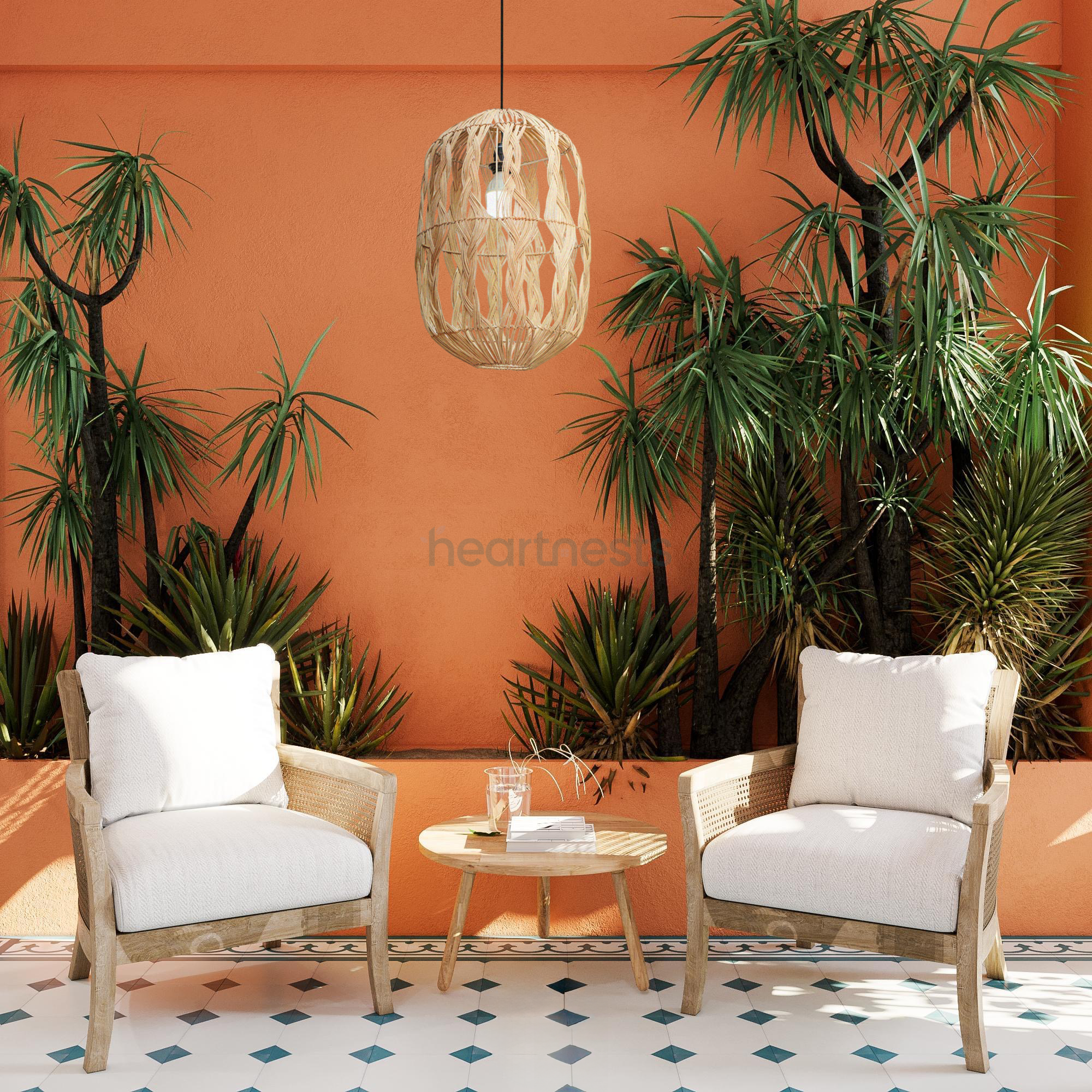 A Heartnests' Malee Rattan Pendant Light is hung above 2 rattan armchairs placed in front of a bright orange colour feature wall and an orange colour retainer with 2 big tropical green trees