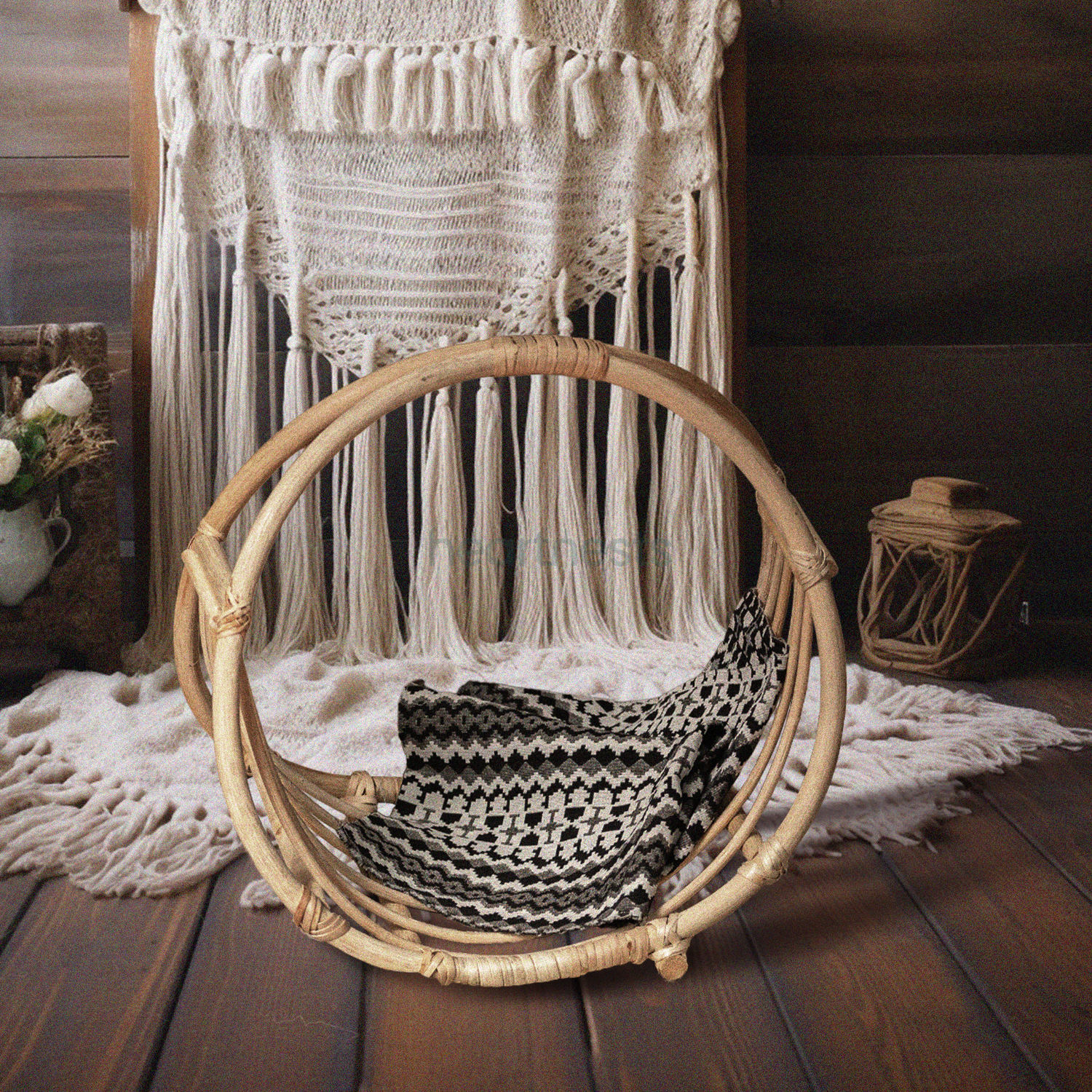 A Heartnests's Wicker Baby Photography Prop is placed on a white macrame and in front of dropping down macrame tassles next to a beautiful white flower vase