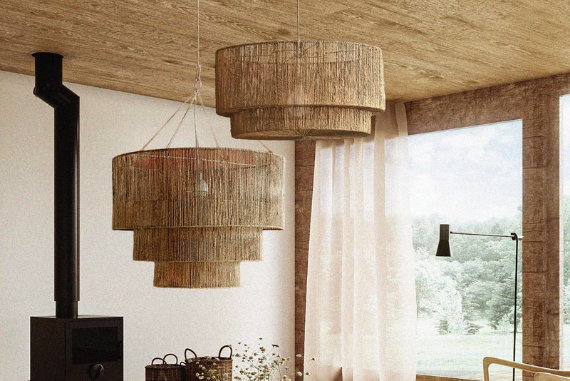 A modern living room interior with two tiered woven natural-fiber pendant lights hanging over a round light wood coffee table. The room features a wood ceiling, a black wood-burning stove, a large window with sheer curtains, and a light wood chair and stool.