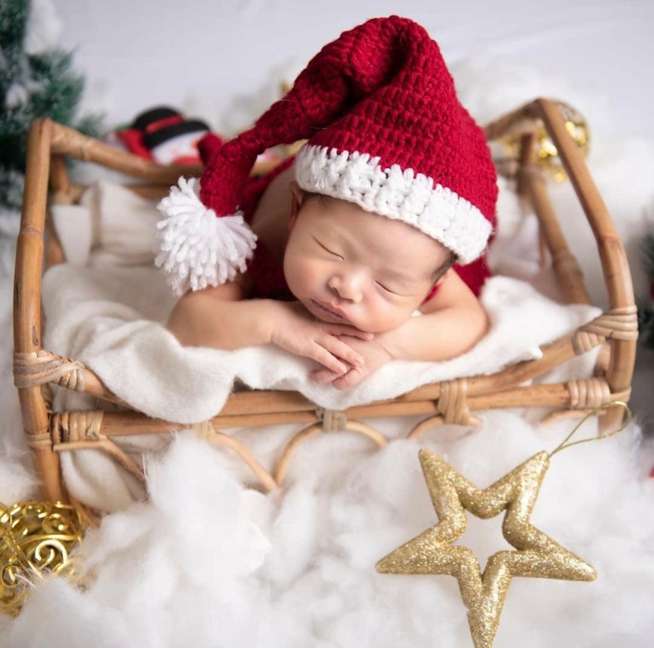 A baby wears red Christmas hat and sleeps so peacefully in Heartnests' Neslia Rattan Baby Prop covered by cloud like background with gold stars