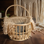 A Heartnests' Kira Rattan Newborn Photography Basket with handles is placed on a dark brown wooden flooring and in front of a white macrame