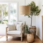 A Heartnests' Lifa Rattan Pendant Light is hung above a 1 gray washed wooden armchair which is next to a pot plant and a seagrass basket in a corner of a living room